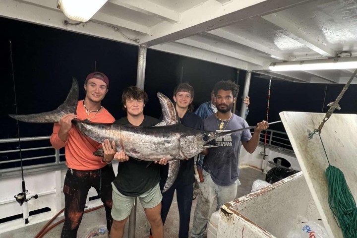 Four people on a boat holding a large fish together.