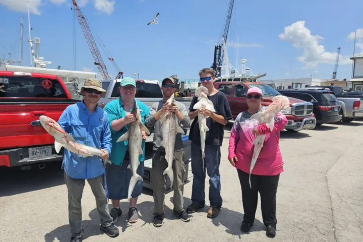 Five people holding fish stand in a parking lot with boats and cranes in the background.