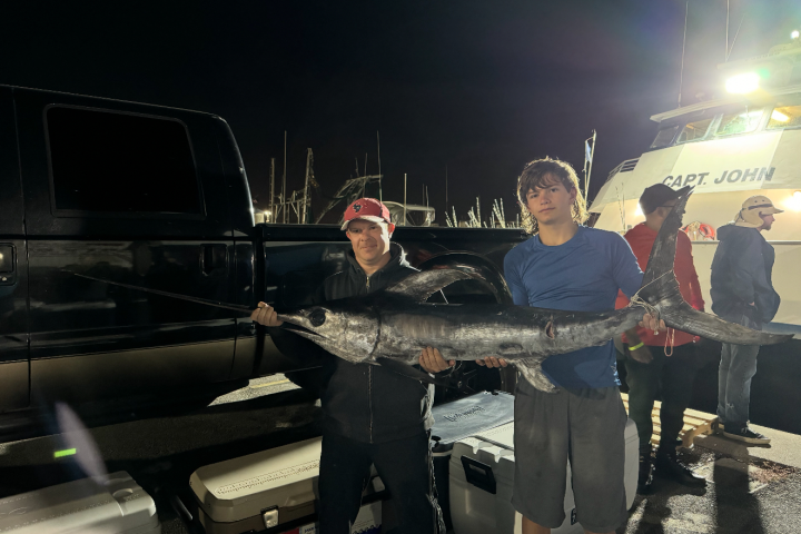 Two people holding a large swordfish at night near a boat and truck.