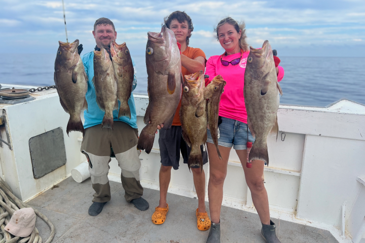 Three people smiling and holding large fish on a boat against a cloudy sky.