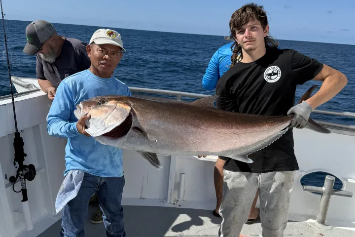 Two people on a boat holding a large fish with ocean in the background.