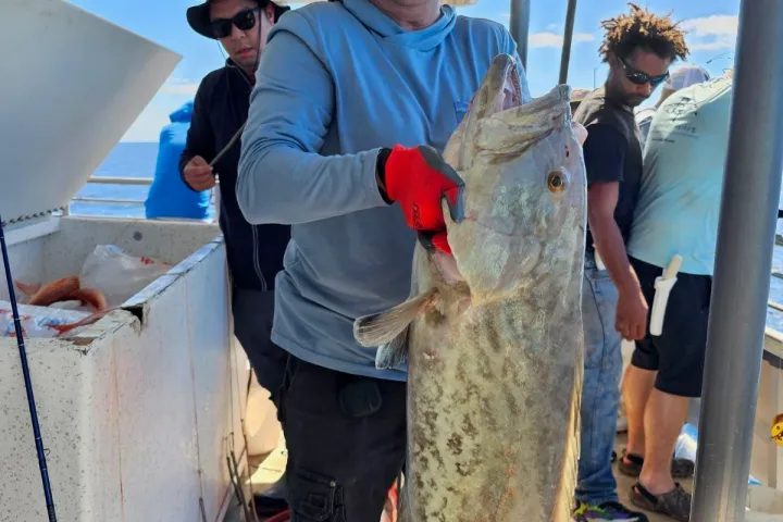 Man in sunglasses holding large fish on a boat with others in the background.